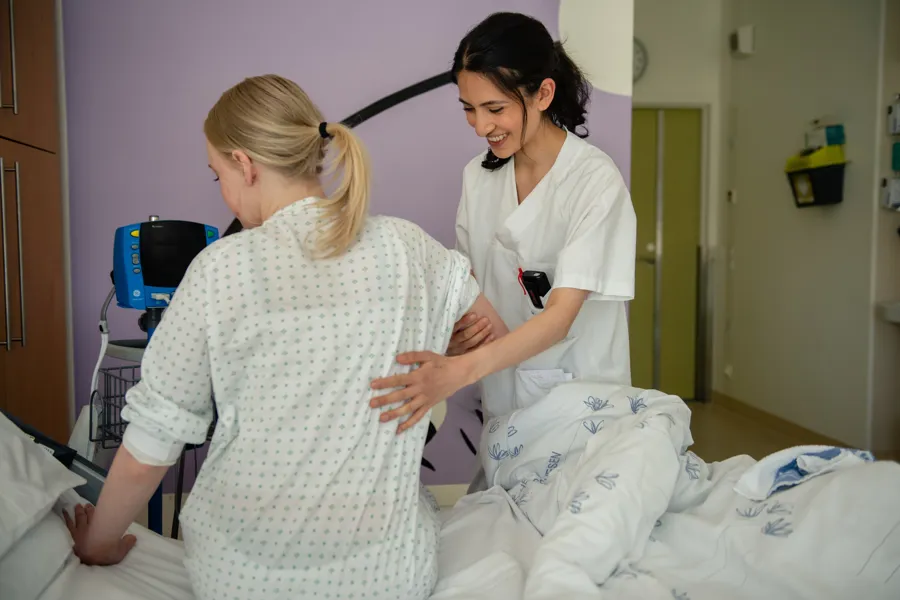 Ung pasient sitter i en seng med ryggen til - en står ved siden av sykepleier holder henne i hånden